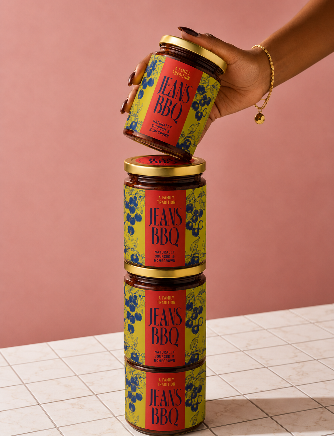 Stack of four jars of Jeans BBQ sauce held by a hand with dark nail polish and a gold bracelet, against a pink background and white tiled surface.