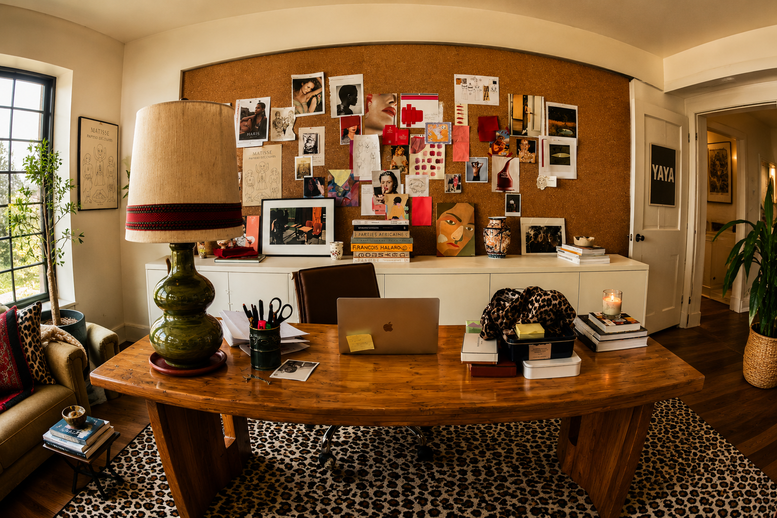 A contemporary home office with a wooden desk, a beige lamp, and various art and photographs pinned on a corkboard. The room features a window with a view of greenery, a leopard print rug, and decorative pillows on an armchair.