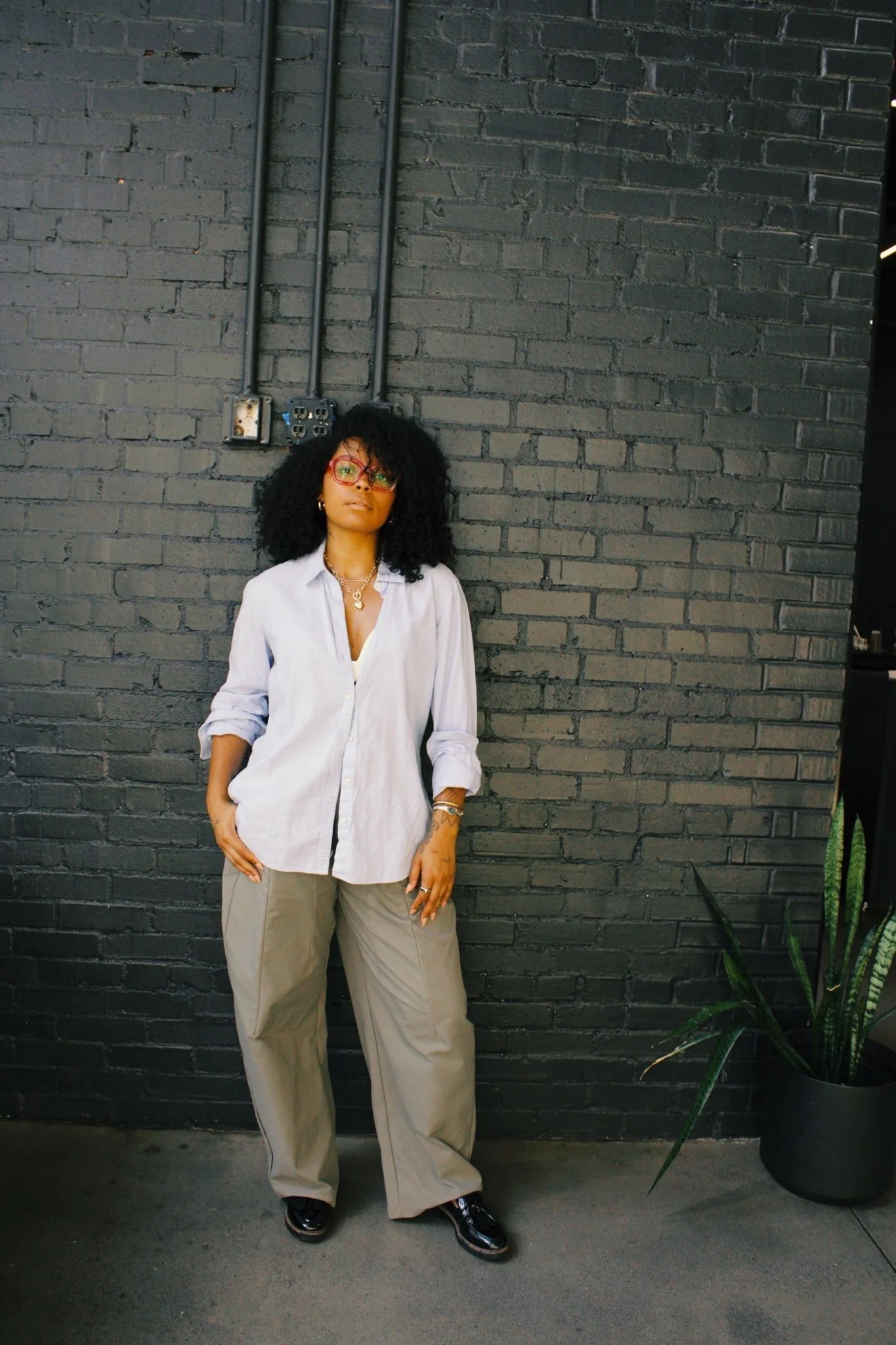 A woman with curly black hair wearing glasses, a white shirt, beige pants, and black shoes standing against a dark gray brick wall with visible electrical outlets and cords. There is a tall green plant in a black pot on the right side of the image.