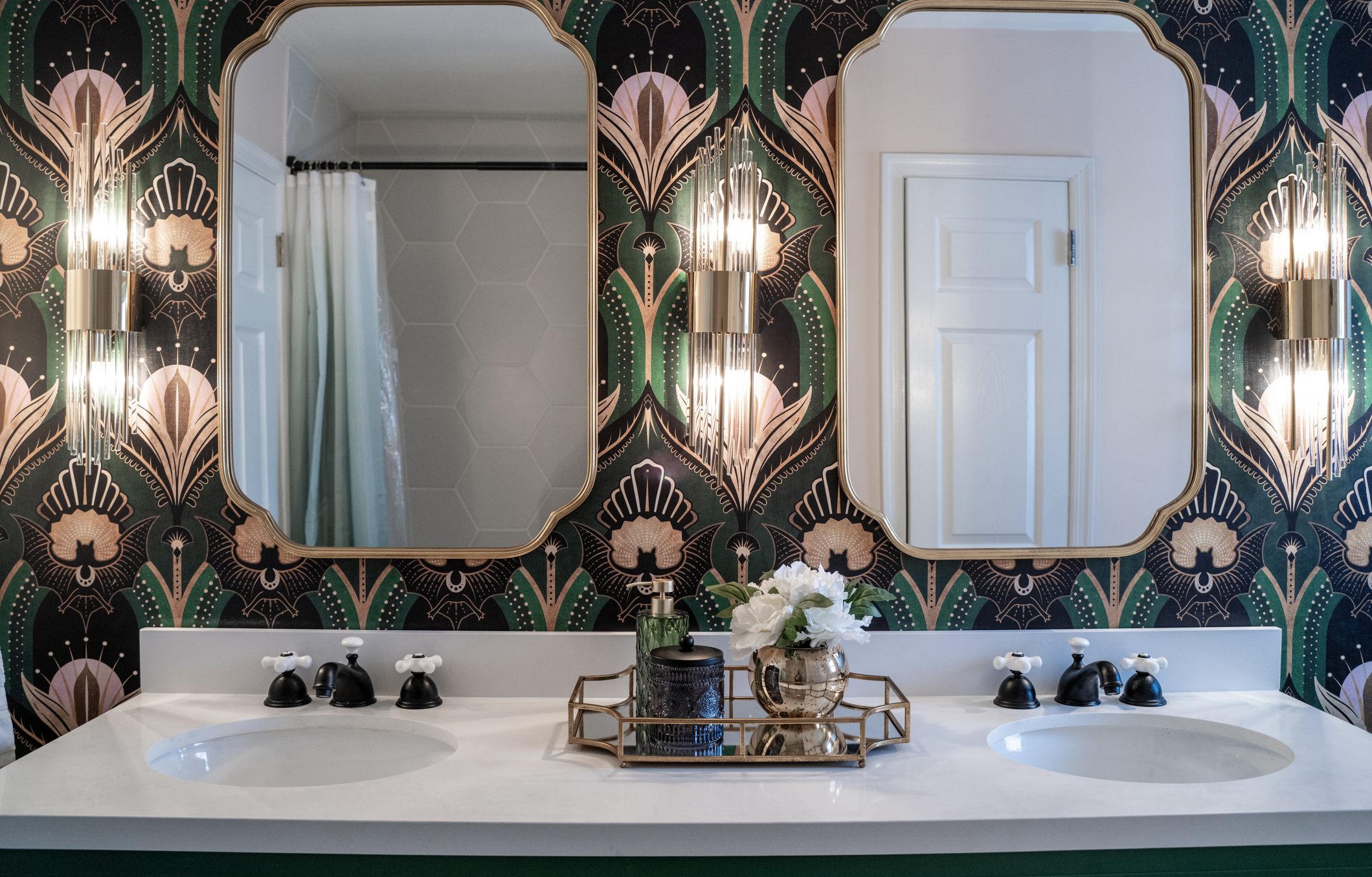 Bathroom double vanity with undermount sinks, black faucets, and a gold tray with decorative items including a green vase, a black container, and a gold vase with white flowers. Two large ornate mirrors and green and black patterned wallpaper with fl