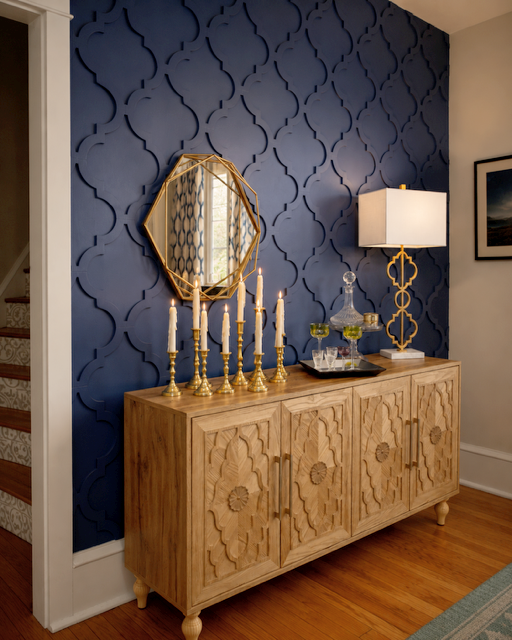 A wooden sideboard with carved floral patterns, a mirror with a gold frame, and candlesticks with lit candles. There is a lamp and decorative items on the sideboard, set against a blue textured wall with a quatrefoil pattern.