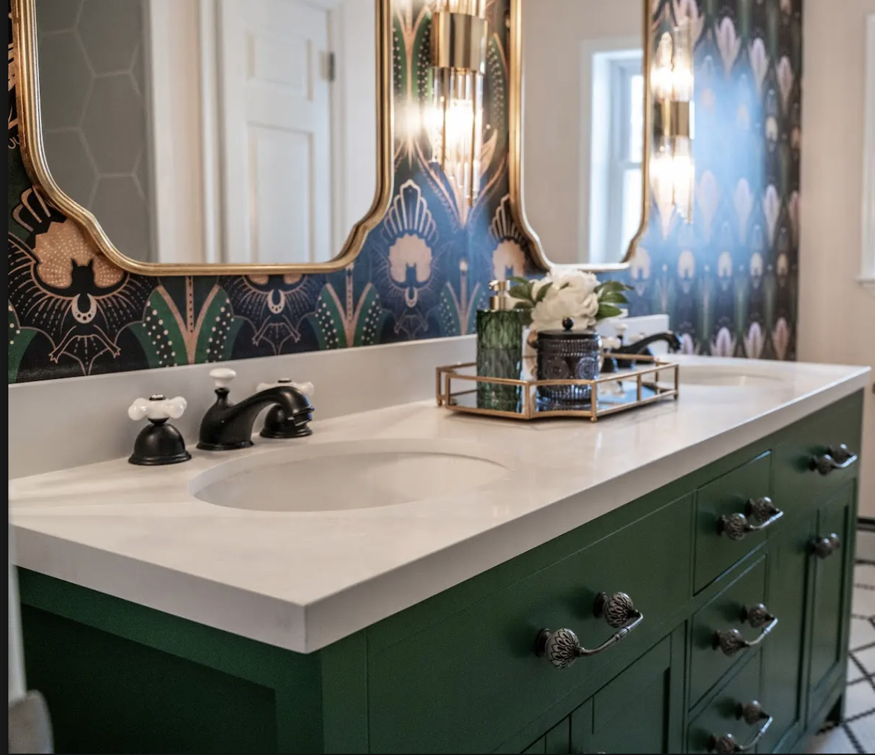 A bathroom vanity with a white countertop, a black faucet, and green cabinetry, decorated with a tray holding a flower arrangement and a vase, standing in front of a patterned wallpaper with framed mirrors and wall sconces.