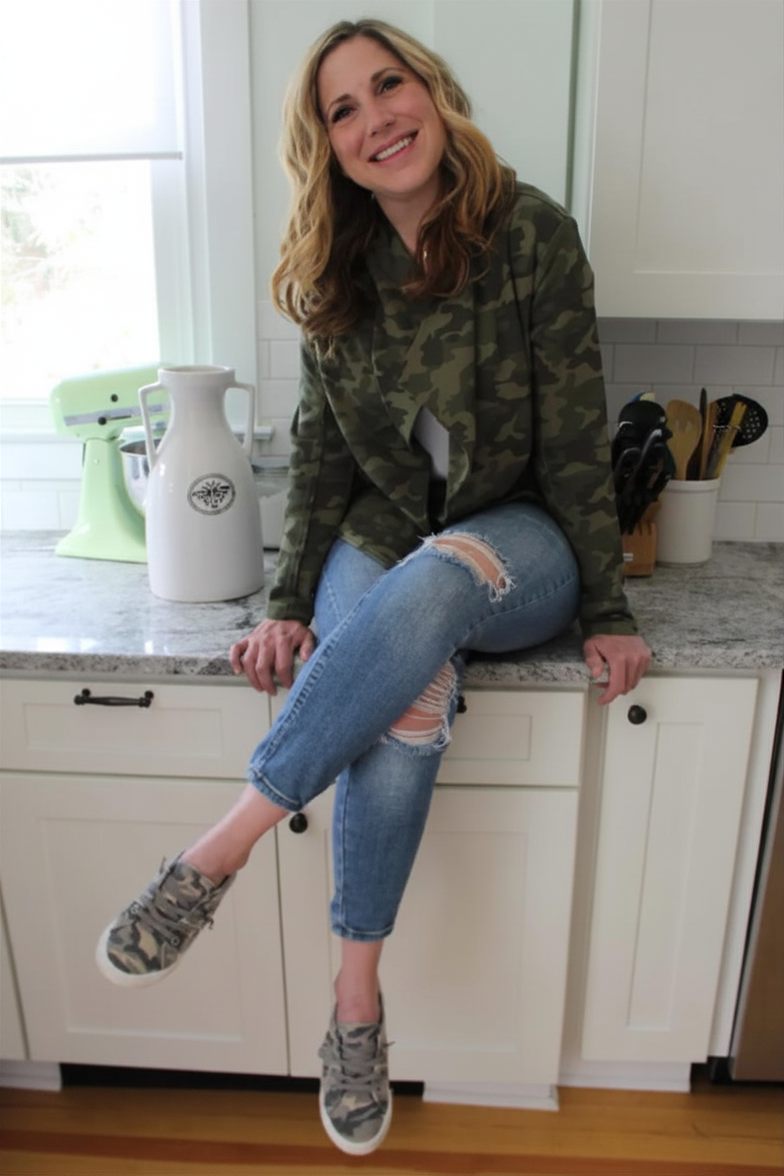A woman with blonde wavy hair wearing a camouflage hoodie, ripped jeans, and camo sneakers sitting on a kitchen counter, smiling at the camera. The kitchen has white cabinets, a gray countertop, and various kitchen utensils and appliances visible in the background.