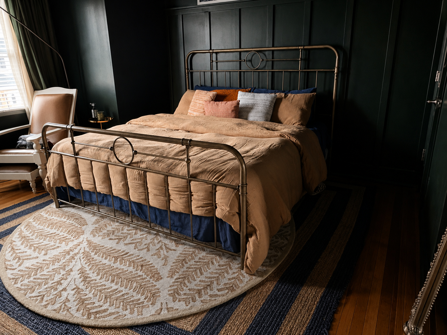 A bedroom with a brass bed featuring beige bedding and decorative pillows, a chair near a window, and a patterned circular rug on wooden floors.