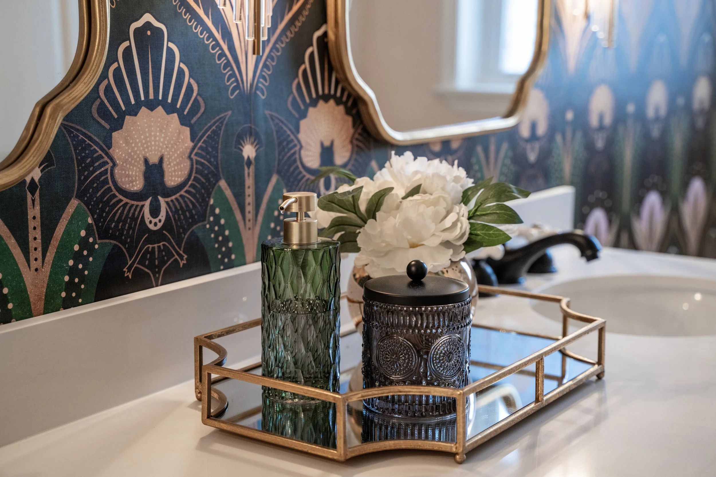 A bathroom countertop with decorative items including a green textured soap dispenser, a black ornate container, and artificial white flowers in a small bowl, all placed on a wooden tray. In the background, there is a mirror with a wooden frame hanging on a wall with a blue and pink patterned wallpaper.