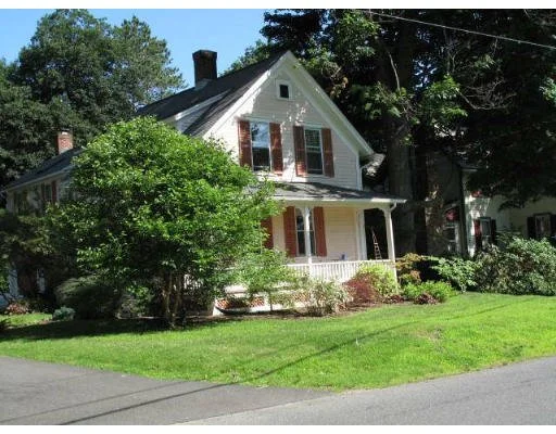 A two-story white house with red shutters, surrounded by greenery, with a lawn and trees in front.