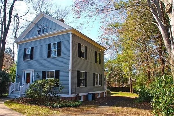 A three-story blue house with black shutters on the windows, a white front door with steps, and surrounded by trees and a lawn.