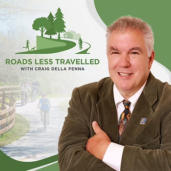 Host with gray hair smiling, standing in front of a promotional graphic for a show called 'Roads Less Travelled' featuring trees, a winding path, and people walking or biking.