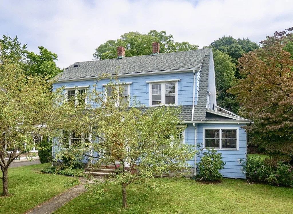 A two-story light blue house with white trim, surrounded by trees and a green lawn, under a cloudy sky.