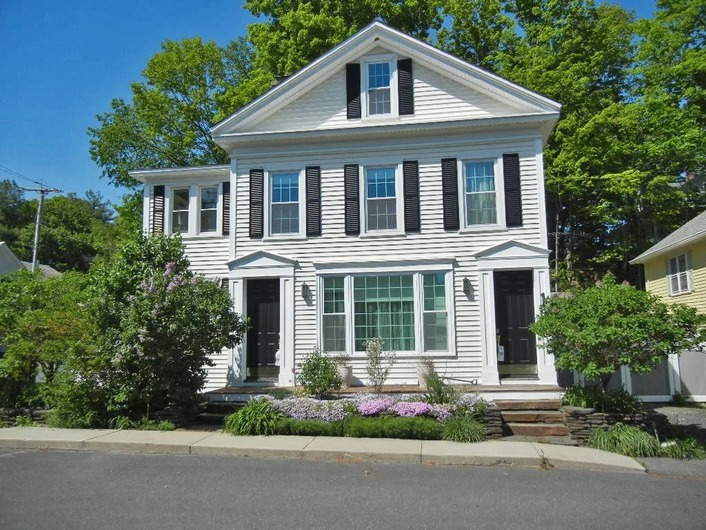 White multi-story house with black shutters, front yard with bushes and flowers, and a sidewalk in front.