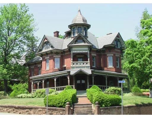 A large, ornate, Victorian-style house with a steep roof, tower, and wraparound porch, surrounded by green trees and bushes.