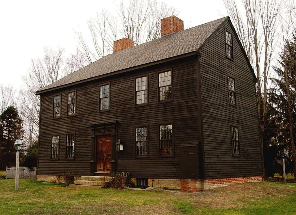 A large three-story house painted black with multiple windows, brick chimneys, and a wooden front door with steps leading up to it, surrounded by a grassy yard and leafless trees.