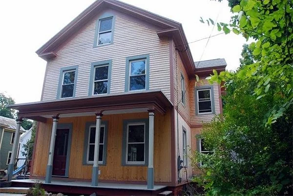 Two-story house under construction, with a front porch supported by four columns, partially finished siding, and surrounded by green trees.