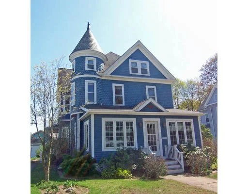 A blue Victorian-style house with a turret and white trim, surrounded by a well-maintained garden and trees.