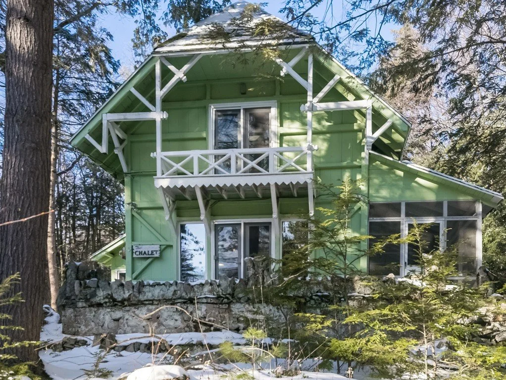 A two-story green house with a sign labeled 'The Chalet' in a wooded area with snow on the ground.
