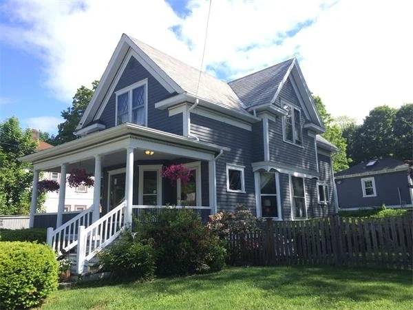 A two-story blue house with white trim, a front porch with white railing, hanging flower baskets, and a well-maintained yard with shrubs and a wooden fence.