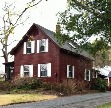 A two-story red house with white window shutters and a black roof, surrounded by trees and bushes, on a grassy lawn with a paved street in front.