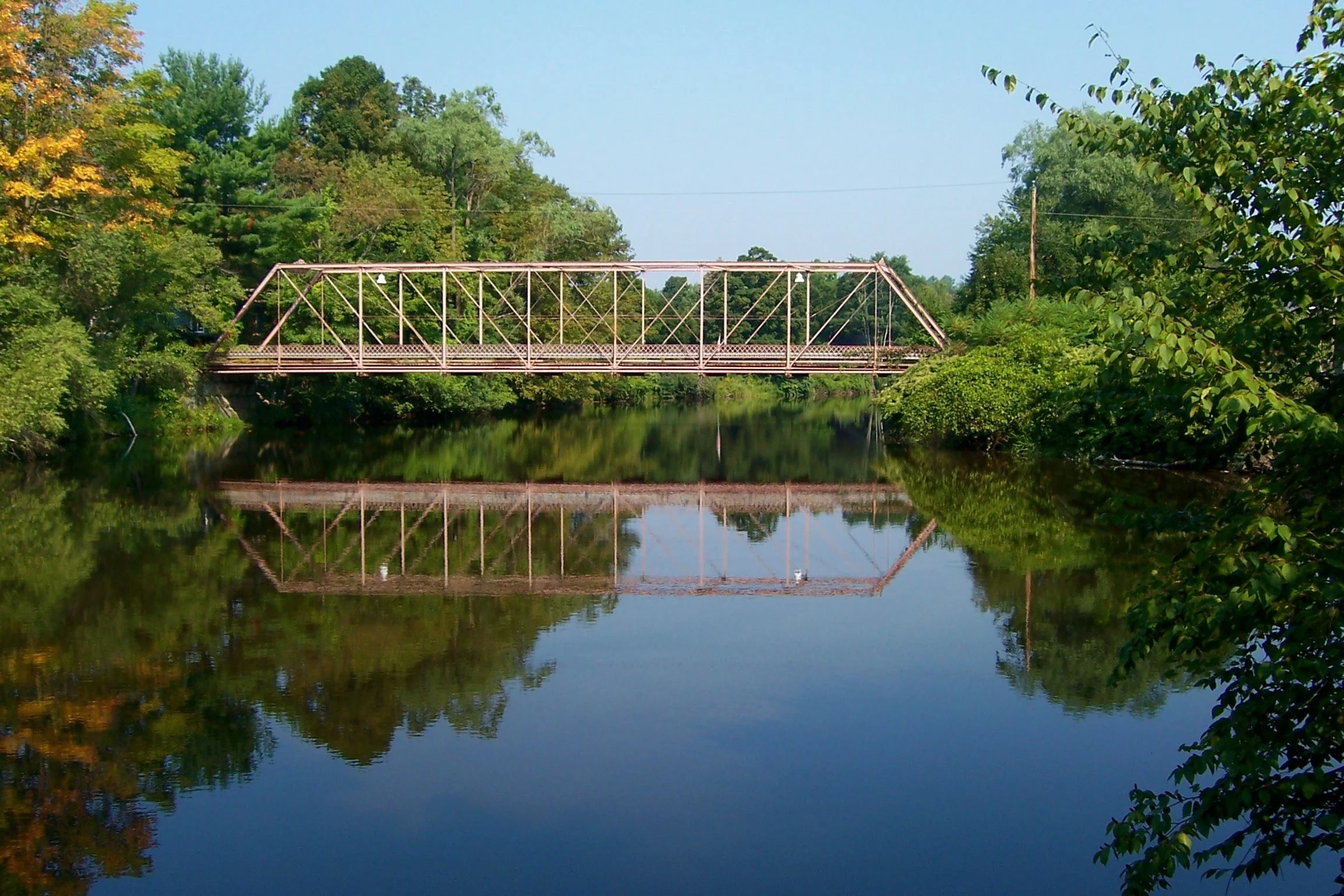 A metal bridge crossing over a calm river, surrounded by lush green trees on a sunny day.