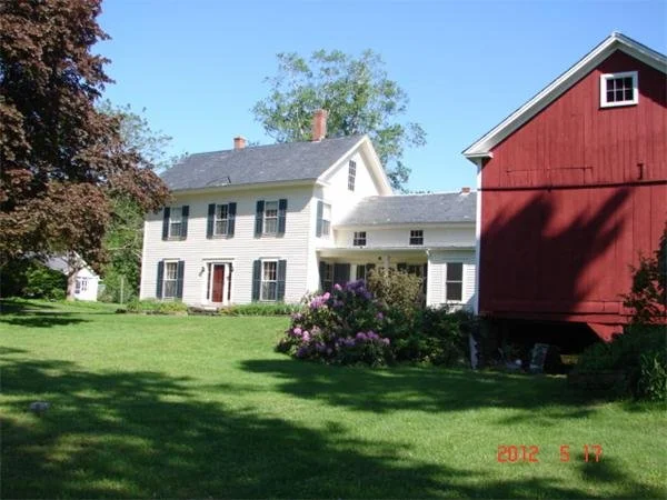 A two-story white house with black shutters and a red barn with a small window under a blue sky.