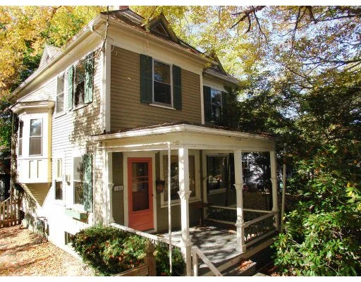A two-story house with beige siding, white trim, green shutters, and a front porch with white columns, surrounded by trees and greenery.