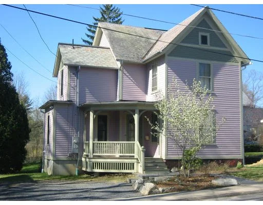 A purple, two-story house with a front porch, located on a residential street with trees and power lines visible.