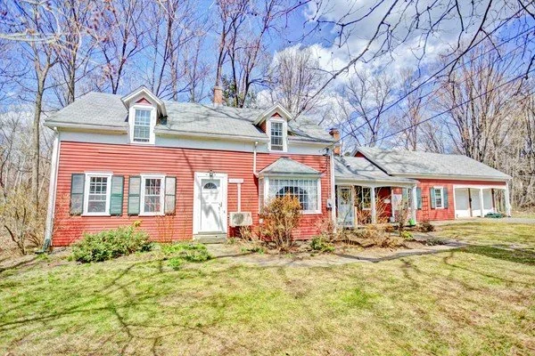 A red two-story house with green shutters, a small front porch, and a large lawn, set in a wooded area with blue sky and leafless trees.