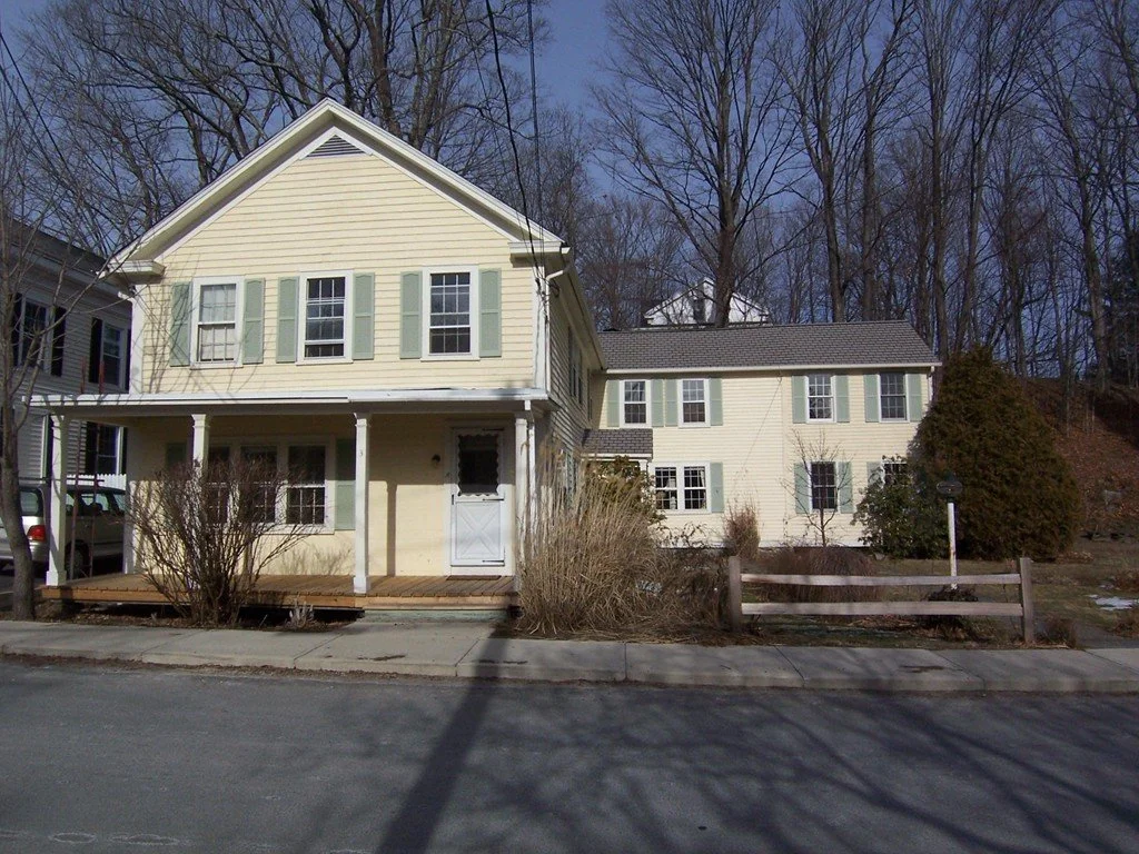 A two-story yellow house with a porch and green shutters, located on a street with leafless trees in the background.