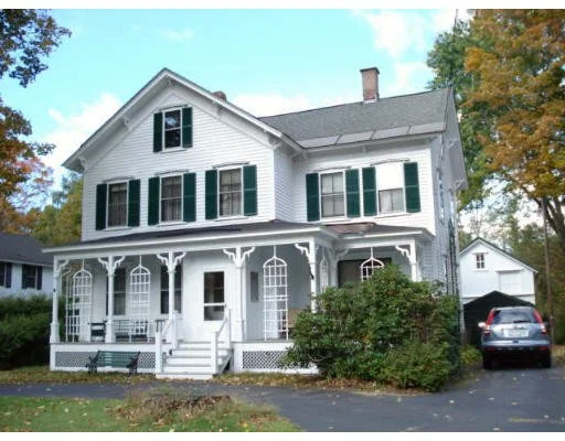 White two-story house with green shutters, a front porch with decorative trim, and a driveway with a parked car, surrounded by trees.
