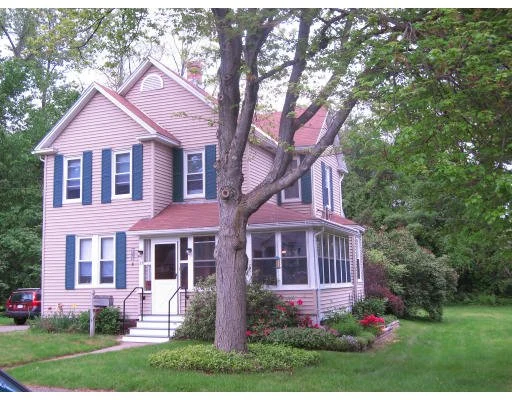 A two-story house with pink siding, blue shutters, and a wraparound porch, surrounded by a green lawn and trees.