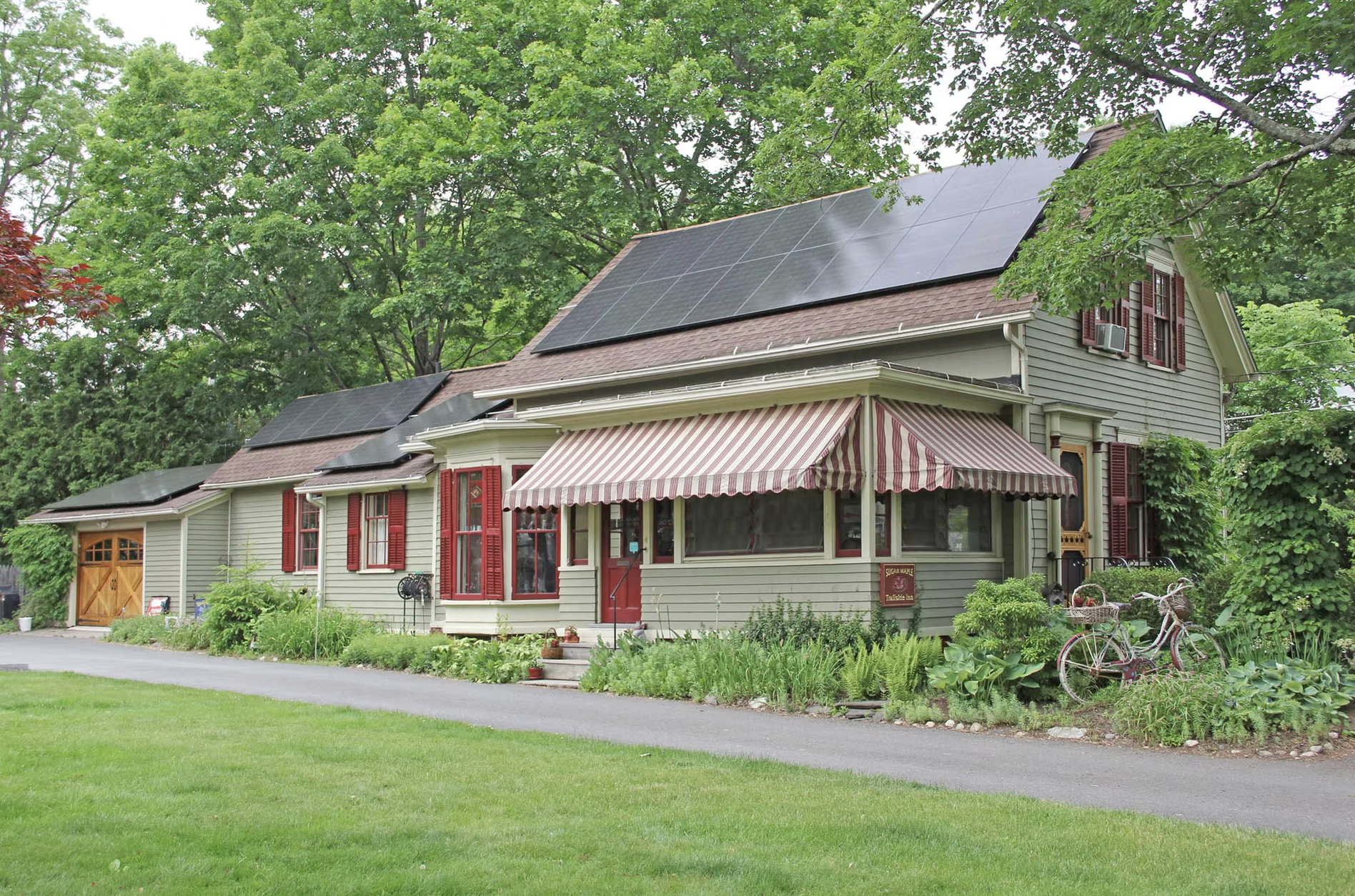 A two-story house with solar panels on the roof and green shutters, surrounded by lush trees and a garden, with bicycles parked outside.