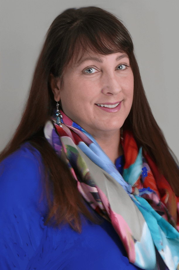 A woman with long brown hair smiling, wearing a blue top and a colorful scarf, against a plain background.