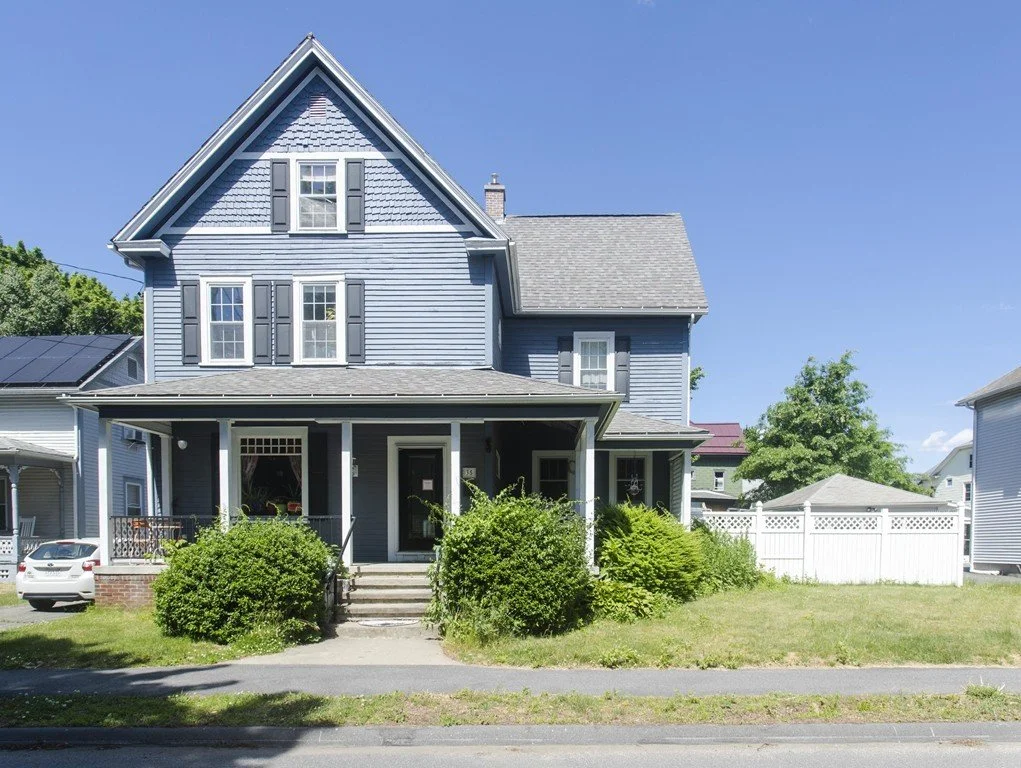 A two-story blue house with gray shutters, a front porch, and a white picket fence in a suburban neighborhood under a clear blue sky.