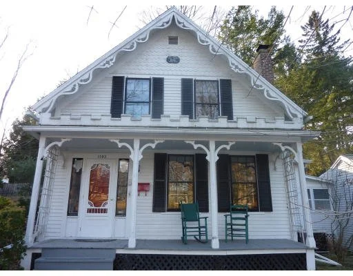 Front view of a white Victorian-style house with decorative trim, black shutters, and a porch with two green chairs.