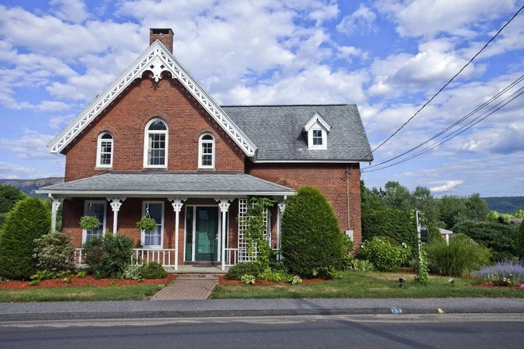A two-story brick house with white trim and a front porch, surrounded by green bushes and plants under a partly cloudy sky.