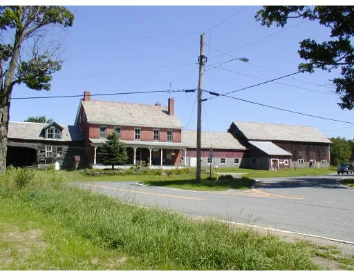 Rural scene with two barns and a house, power lines, and a paved road in the foreground, surrounded by green grass and trees.