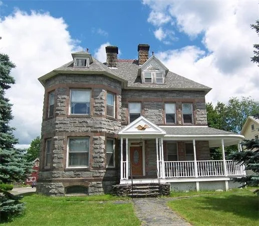 A large, historic stone house with a porch and multiple windows, surrounded by a lawn and trees, under a partly cloudy sky.