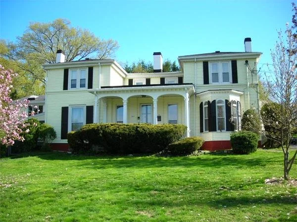 A large, two-story house with yellow siding, black shutters, and a red foundation, surrounded by green grass and trees.