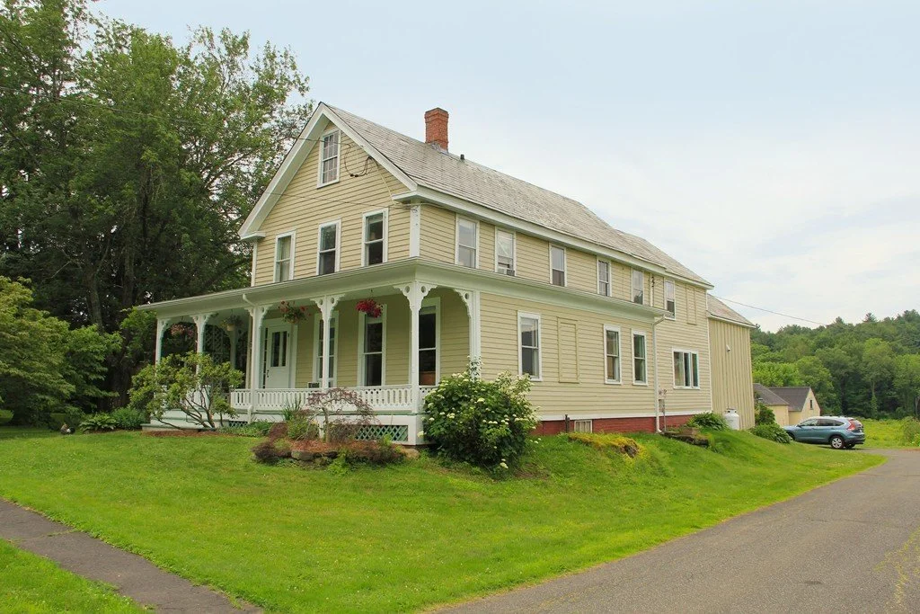 A large, vintage yellow house with white trim and a wrap-around porch, surrounded by green lawn and trees in the background.