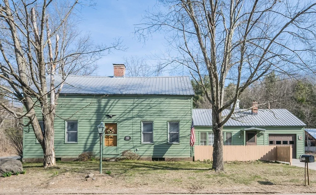 A green house with a metal roof, two leafless trees in front, an American flag, and a mailbox.