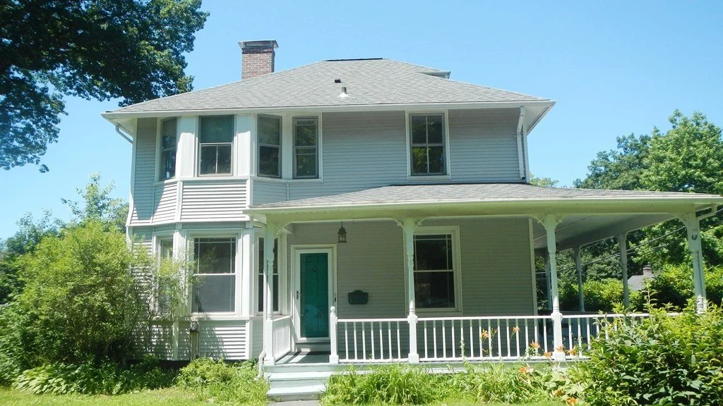 A two-story house with light gray siding, a covered front porch with white railings, numerous windows, and a chimney, surrounded by green trees and bushes under a bright blue sky.