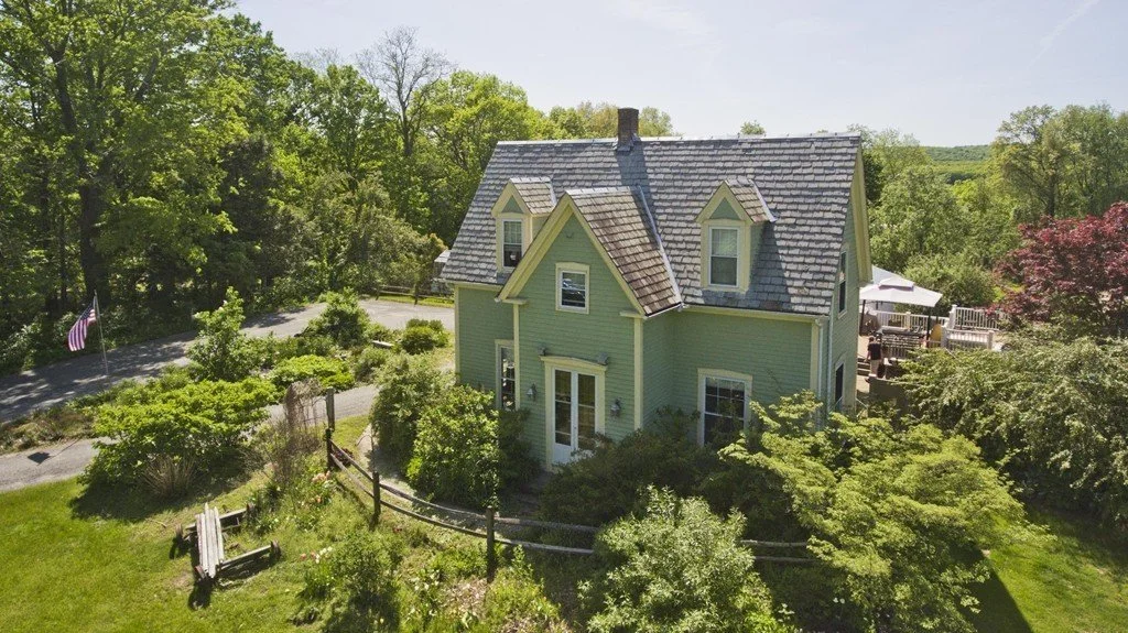 An aerial view of a light green house with a gray roof surrounded by trees, shrubs, and a grassy yard, with a driveway and an American flag in the background.