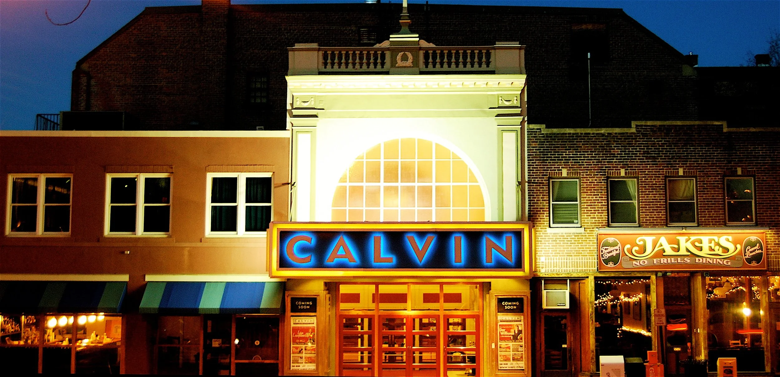 Nighttime exterior view of a building with illuminated signs for Calvin and Jake's restaurant, featuring a large lit Sign with blue letters reading 'CALVIN' and a Coca-Cola themed sign for Jakes restaurant, with a large decorative window above the signs.