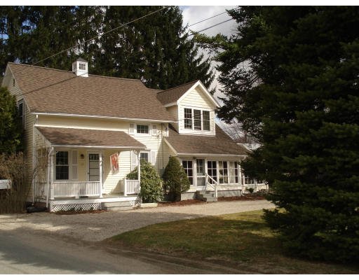 A large white house with a porch, multiple windows, and a brown roof, surrounded by trees and situated on a grassy area.