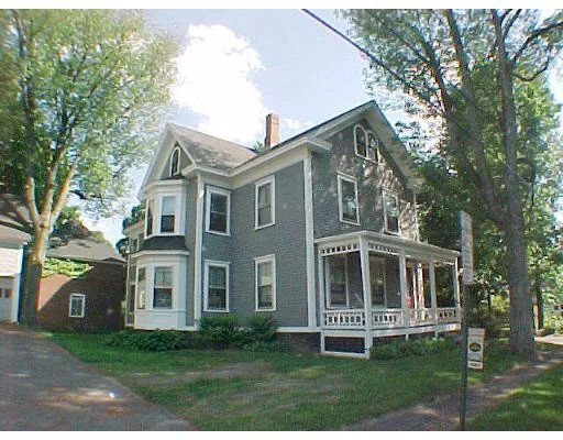 A gray, three-story Victorian house with a front porch, multiple windows, surrounded by trees and grass.