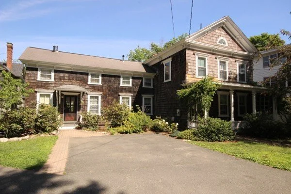A large two-story house with dark brown wooden siding, brick accents, multiple windows, and a front porch, surrounded by trees and shrubs, with a driveway in front.