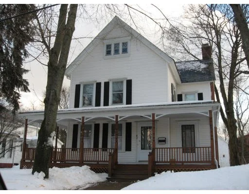 A white two-story house with black shutters, a gabled roof, and a front porch surrounded by snow.