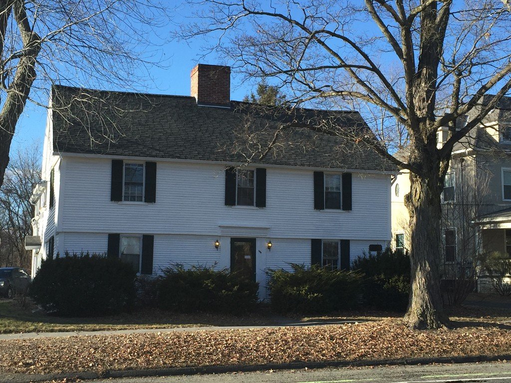White two-story house with black shutters, leafless trees, and a front yard with bushes on a sunny day