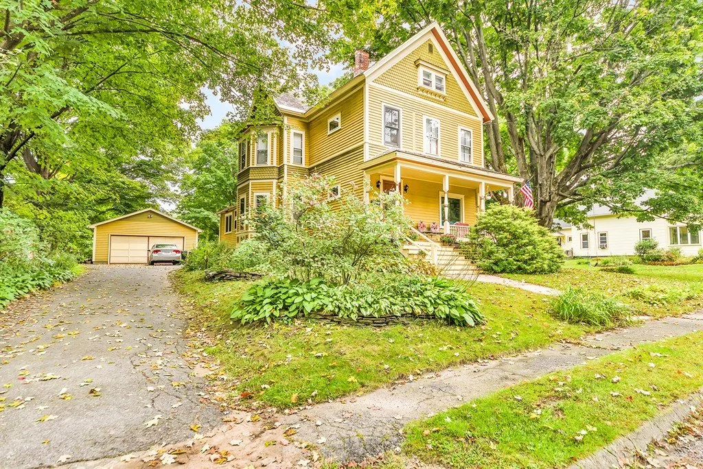 Yellow Victorian-style house with a front porch, surrounded by green trees and lawn, with a detached garage and a driveway.