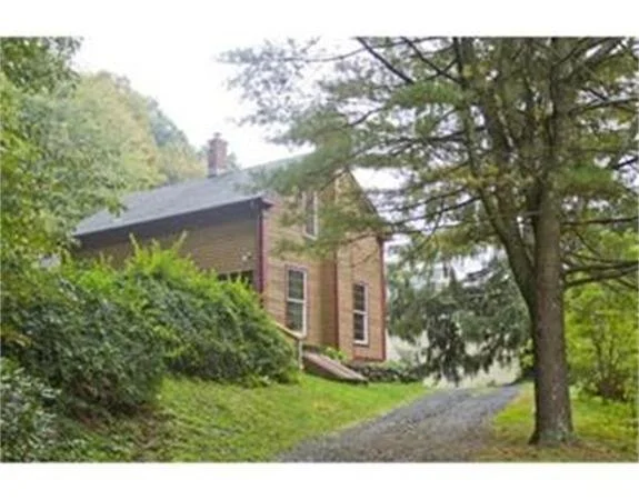 A brick house surrounded by green trees and bushes, with a gravel pathway leading to it.