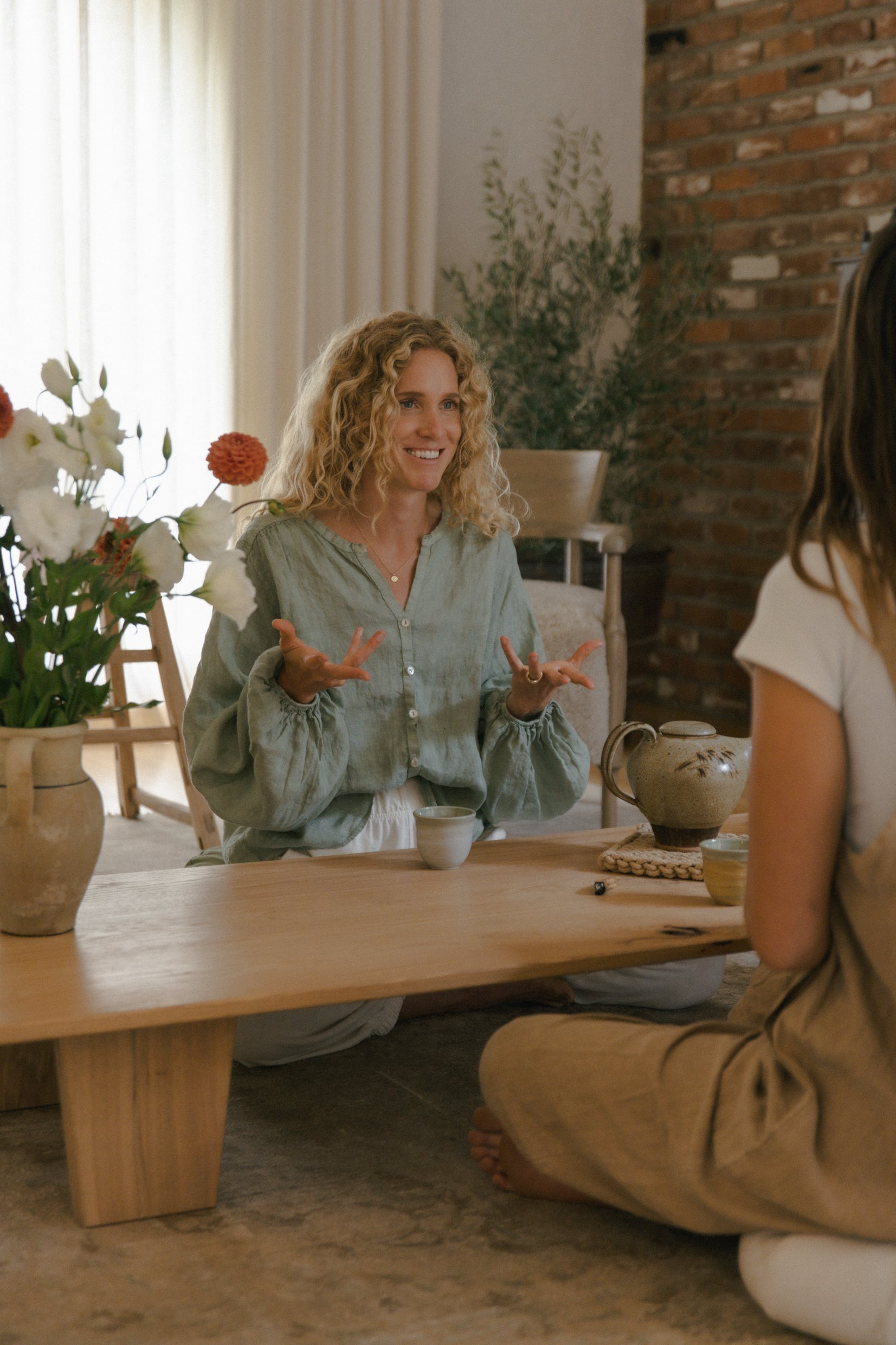 A woman with curly blonde hair, wearing a light green blouse, is sitting at a wooden table, smiling and gesturing with her hands during a conversation with another person. The setting is a cozy room with a brick wall, a large window with curtains, and plants. There are flowers in vases on the table.